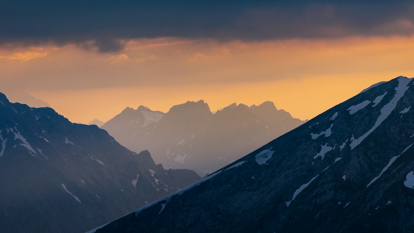 Schneebedeckte Berggipfel bei Sonnenuntergang vor einem orange-gelben Himmel.